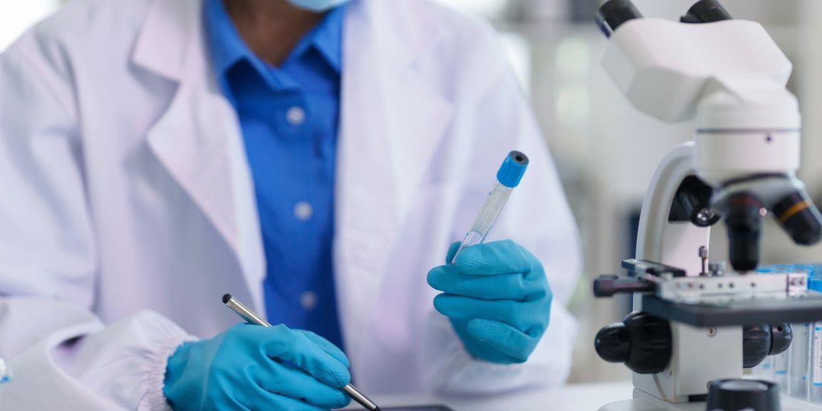 A scientist in a lab coat and gloves examines a sample under a microscope in a laboratory setting.
