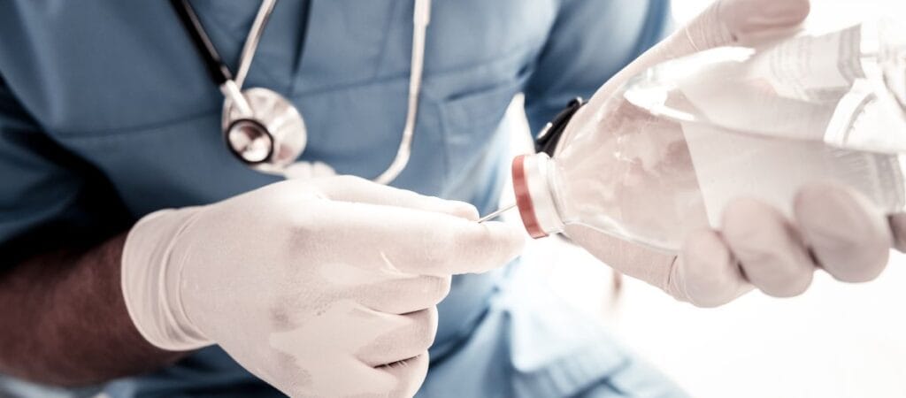 A doctor in a white coat holds a clear bottle of water, smiling and looking at the camera in a clinical setting.