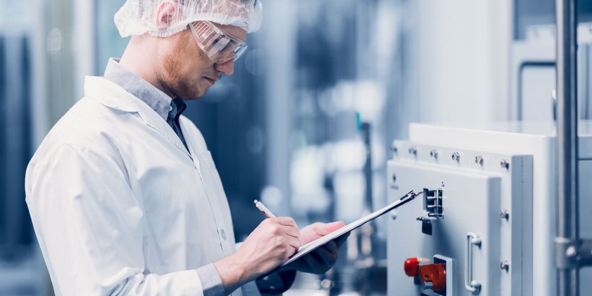 A man in a lab coat is writing notes on a clipboard, focused on his task in a laboratory setting.