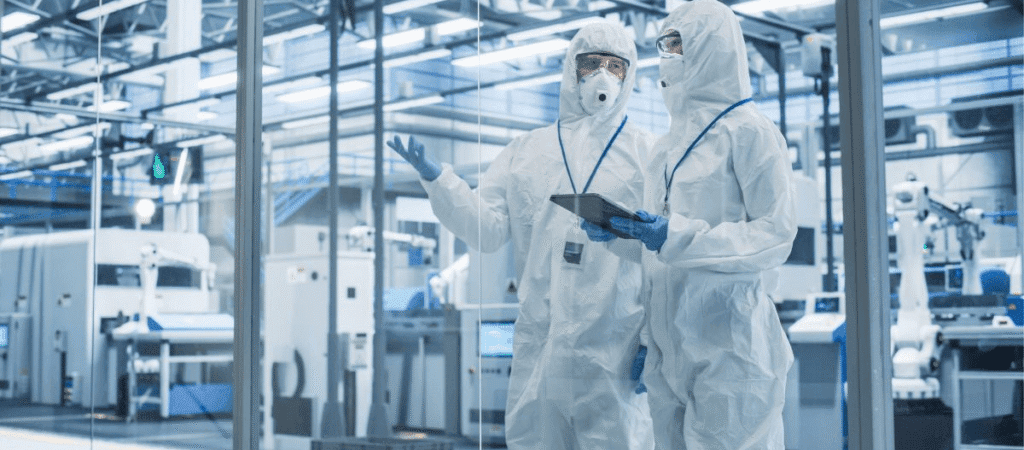 Two individuals in protective suits stand inside a factory, surrounded by industrial equipment and machinery.